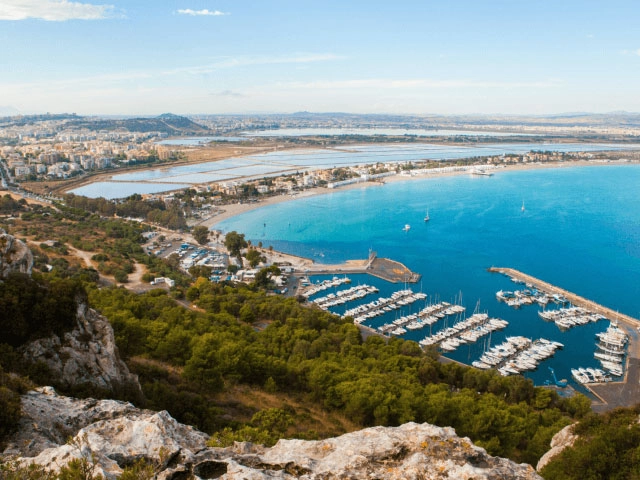 Nell'immagine, una vista panoramica del mare: si vede un porticciolo con delle barche e, in primissimo piano, un promontorio con della boscaglia.